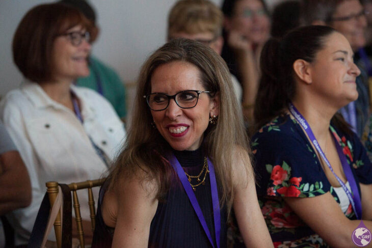 A woman with glanzmann's thrombasthenia wearing glasses and purple lanyard sitting at a conference.