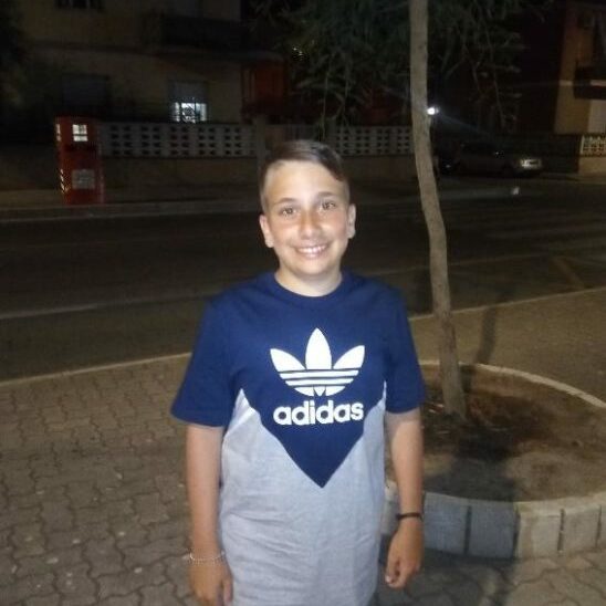 A young boy wearing an adidas t-shirt standing on a sidewalk at night with glanzmann's thrombasthenia.